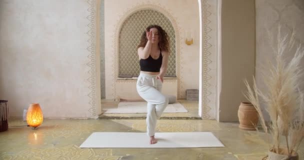 Спортивная кудрявая женщина занимается упражнениями по йоге. Стройная девушка занимается йогой в яркой, атмосферной комнате для йоги утром — стоковое видео