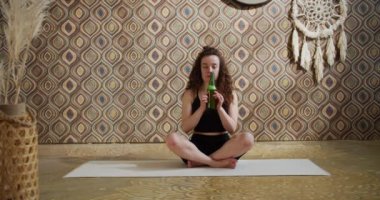 Bira yogası. Kıvırcık saçlı genç bir kadın yoga yapıyor ve bir şişe birayla meditasyon yapıyor. Atletik ince kız sabahları parlak, atmosferik yoga odasında bira yogası yapıyor.
