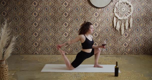 Wijn yoga. Curly-Hired Jonge vrouw doet yoga stretching oefening met een glas wijn. Sportief slank meisje doet wijn yoga in een heldere, atmosferische yoga kamer in de ochtend — Stockvideo