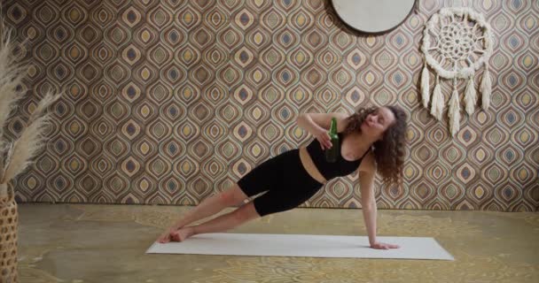 Öl yoga. Curly-Haired Ung kvinna som gör yoga stretching övning med en flaska öl. Sportig smal tjej som gör ölyoga i ett ljust, stämningsfullt yogarum på morgonen — Stockvideo