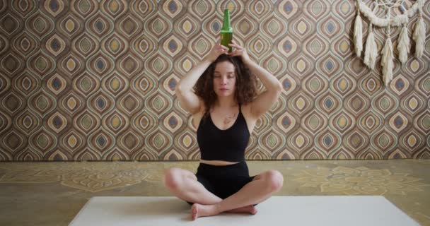 Yoga de la cerveza. Una joven de pelo rizado haciendo un ejercicio de yoga y meditando con una botella de cerveza en la cabeza. Atlética chica delgada hace yoga cerveza en una sala de yoga luminosa y atmosférica por la mañana — Vídeo de stock