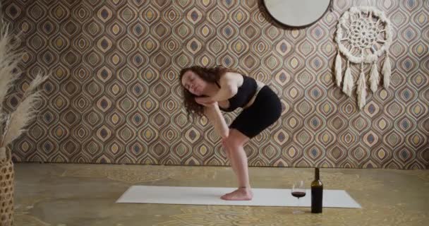 Vinyoga. Curly-Haired Ung kvinna som gör yoga stretching övning med ett glas vin. Sportig smal tjej som gör vinyoga i ett ljust, stämningsfullt yogarum på morgonen — Stockvideo