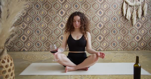 Yoga du vin. Une jeune femme aux cheveux bouclés qui fait un exercice de yoga et médite avec un verre de vin. Athlétique fille mince fait du yoga vin dans une salle de yoga lumineux et atmosphérique le matin — Video