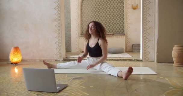 Giovane donna dai capelli ricci istruttore di yoga coaching online, facendo video di lezioni di yoga sulla fotocamera del computer portatile. Attraente ragazza in forma guardando lezione di yoga nel computer portatile. — Video Stock