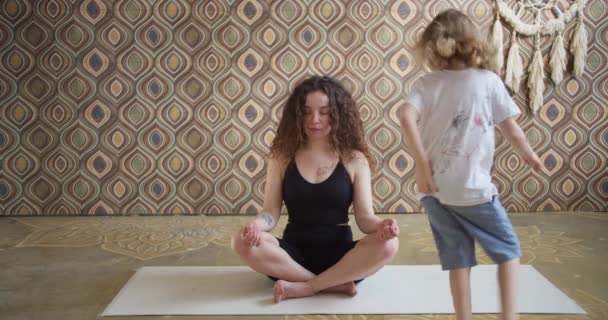 Mère aux cheveux bouclés médite tout en étant assis sur un tapis de yoga, tandis que la fille de bébé énergique active saute tout en jouant, calme jeune maman fait des exercices de yoga à la maison pour soulager le stress tout en se relaxant avec un — Video