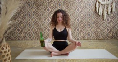 Bira yogası. Kıvırcık saçlı genç bir kadın yoga yapıyor ve bir şişe birayla meditasyon yapıyor. Atletik ince kız sabahları parlak, atmosferik yoga odasında bira yogası yapıyor.