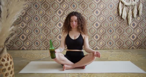 啤酒瑜伽。一位卷曲头发的年轻女子正在做瑜伽运动，一边喝着一瓶啤酒，一边沉思。运动健美的女孩早上在明亮的氛围中做啤酒瑜伽 — 图库视频影像