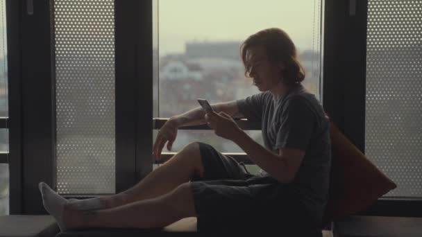 Bonito hipster homem senta-se sozinho na frente de uma janela panorâmica com um smartphone. Aprendizagem online, trabalho doméstico, freelance. Vírus da Corona - ficar e trabalhar em casa auto-isolamento, auto-quarentena — Vídeo de Stock