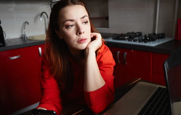 Distracted from work worried young freelancer woman sitting on kitchen with laptop, thinking of problems