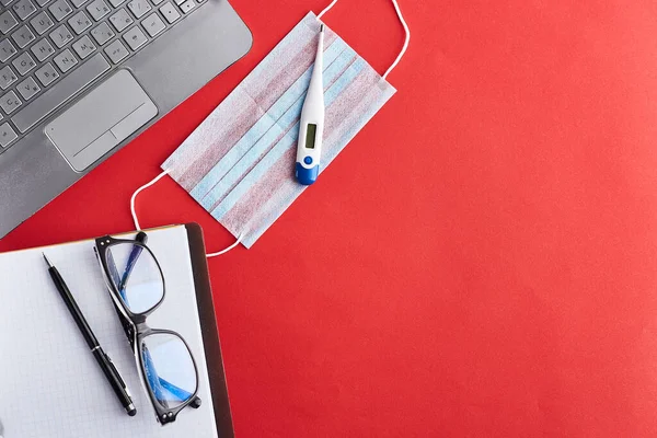 Workplace at quarantine. Home working workplace. Remote work from home. Blank white paper and black pen, medical mask, thermometer, headphones and laptop on red background