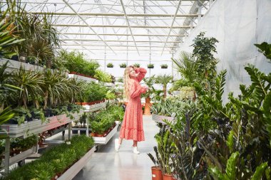 Kırmızı elbiseli genç sarışın kadın bir seranın içinde dans ediyor. Saksıda çiçekler olan sanayi serası.