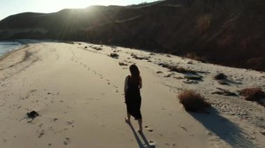 Kumsalda yürüyen uzun siyah elbiseli kızıl saçlı kadın. Kumsalda şehvetli bir yürüyüş. Kıyı şeridi boyunca uçurumları olan kumlu sahil manzarası.