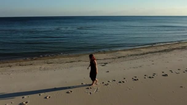 穿着黑色长裙的红头发女人走在海滩上 性感地走在沙滩上 海岸线上有悬崖的沙滩的空中景观 — 图库视频影像