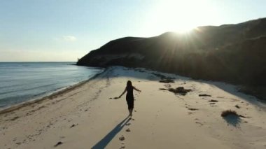Kumsalda yürüyen uzun siyah elbiseli kızıl saçlı kadın. Kumsalda şehvetli bir yürüyüş. Kıyı şeridi boyunca uçurumları olan kumlu sahil manzarası.
