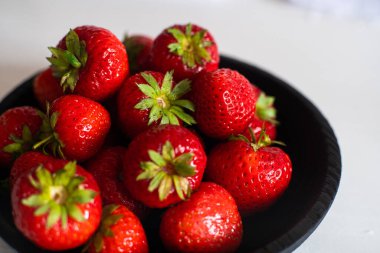 Taze, olgun çileklerle dolu siyah bir kase temiz beyaz bir masada duruyor.