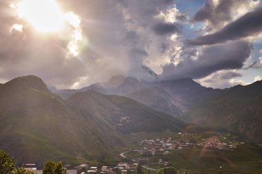 Georgia mı? Kazbek Dağı Zirvesi. Bulutlu bir akşamda Kafkasya bölgesinin aşağısındaki köyle birlikte dağ manzarasının üzerinde gün batımı. Stepantsminda, Georgia. Sakartvelo