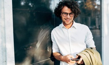 Açık havada gülümseyerek poz veren göz giysili bir iş adamı. Erkek girişimci şehir caddesinde dinleniyor. Sıradan giyinen zeki adam, işten sonra dışarıda yürüyen kıvırcık saçlı gözlük takıyor.