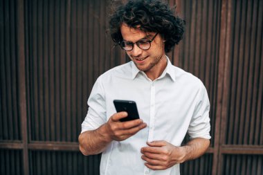 Gözlüklü, beyaz gömlekli mutlu iş adamının cep telefonundan mesajlaşması. Yakışıklı kıvırcık erkek dışarıda bir iş arkadaşıyla telefon üzerinden online konferans yapıyor..