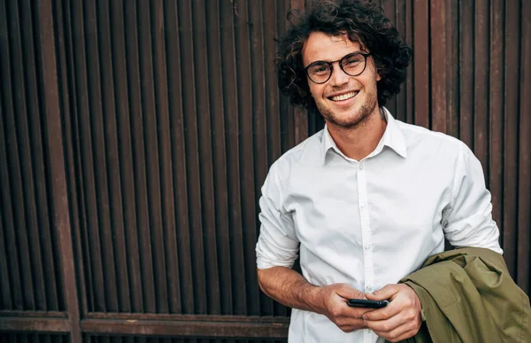 Açık havada gülümseyerek gülümseyen başarılı bir iş adamı. Cep telefonundan mutlu erkek girişimci mesajları. Sıradan giyinen zeki adam, işten sonra dışarıda yürüyen kıvırcık saçlı gözlük takıyor.