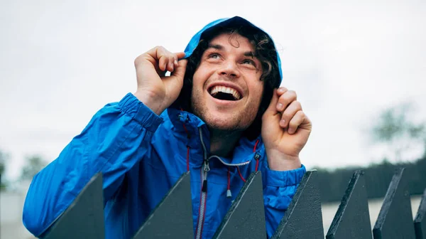 Joyful man wearing blue raincoat during the rain. Happy male in blue raincoat enjoying the rain next to the house. Young man has a joyful expression and looking up tp the sky.