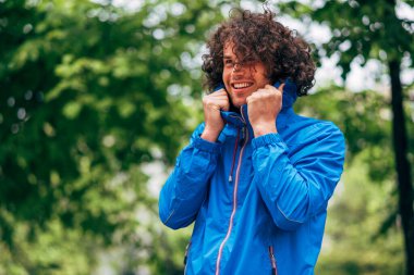Dışarıda yağmurda mavi yağmurluk giyen kıvırcık saçlı, gülümseyen adam. Mavi yağmurluklu olumlu erkek şehir sokaklarında yağmurun tadını çıkarıyor. Yağmurlu havada insanın neşeli bir ifadesi vardır..