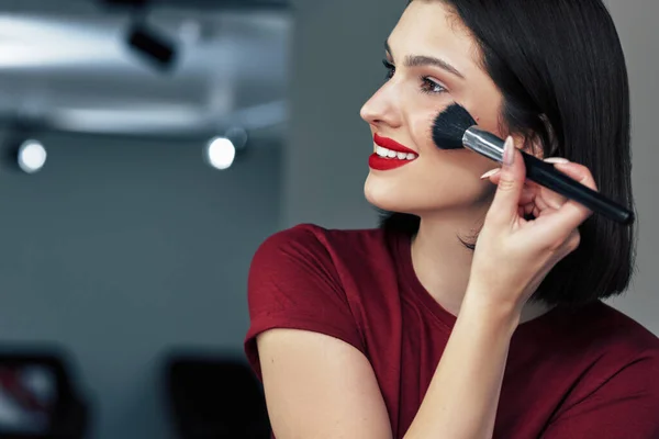 Side view portrait of a gorgeous brunette girl with red lips making makeup tutorial during online lessons. Pretty young woman making herself makeup with organic cosmetics.