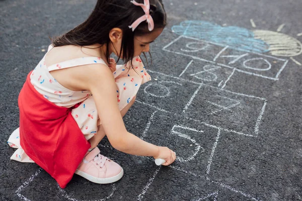 A cute little girl drawing with chalk hopscotch on the playground. Child playing the game outside. The kid wears a dress during drawing on the pavement. Activities and games for children outside.