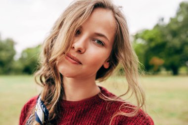 Yakın plan, bordo kazaklı güzel bir genç kadının doğal arka planda poz veren kameraya doğrudan baktığı samimi bir portresi.. 