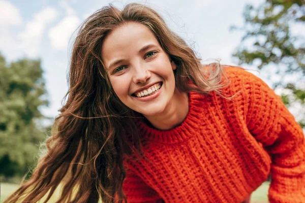 Candid portrait of a cheerful young woman wearing an orange sweater spending time in the park. The pretty female has a joyful expression, resting outdoors on nature background.