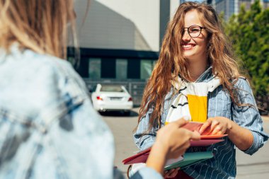 Üniversite kampüsünde açık havada kitapları paylaşan iki öğrenci. Genç kız üniversite öğrencileri şehir sokaklarında klasörleri ve kitapları paylaşıyor..
