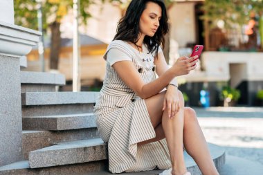 Şehir caddesinde telefon kullanan esmer bir iş kadınının yatay görüntüsü. Kadın girişimci elinde cep telefonuyla iş arkadaşlarını bekliyor..