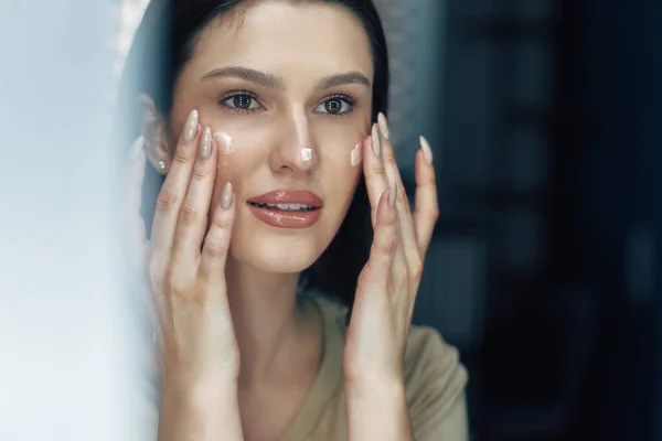 Candid portrait of beautiful woman applying cream on her face. Closeup of a brunette young woman using moisturizing cream and looking to the mirror.