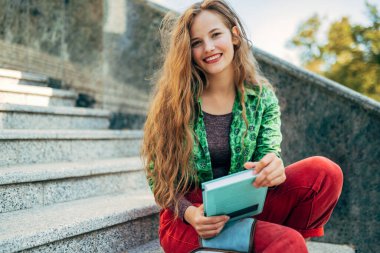 Güneşli bir günde kampüsün merdivenlerinde oturmuş kitap tutan güzel bir kız öğrenci. Sıradan giyinmiş neşeli genç bir kadın dışarıda dinleniyor..