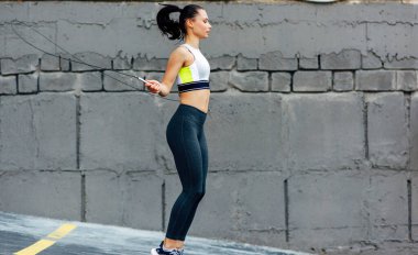 Kararlı ve zinde bir kadının beton duvarın yanında ip atlarken tam yatay çekim. Sporcu kadın dışarıda ip atlamakla egzersiz yapıyor..