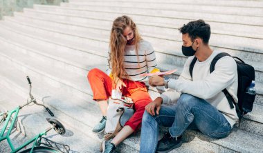 Koruyucu maskeli iki genç öğrenci dışarıda üniversite kampüsünün merdivenlerinde oturmuş konuşuyorlar. Genç bir kadın ve genç bir adam takım çalışması için üniversitenin merdivenlerinde oturuyorlar..