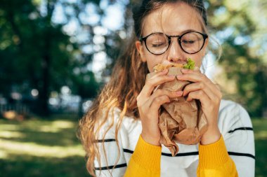 Güzel bir kız öğrencinin dışarıda bir sandviçle öğle yemeği yemesi. Gözlüklü sarışın genç bir kadın parkta fast food yemeye ara veriyor..