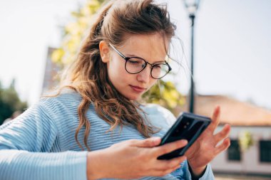 Mavi kazaklı, gözlüklü, güzel bir kadın güneşli bir günde bankta oturmuş açık havada akıllı telefonlardan haber okuyor. Mutlu kadın internette konuşurken dışarıda cep telefonuyla dinleniyor..