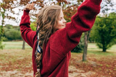 Kolları açık gülümseyen, arka planda dans eden, özgür hisseden genç bir kadın. Parkta eğlenen mutlu bir kadın. Ruh sağlığı konsepti