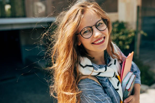 Candid portrait of a happy young student woman with long red hair smiling and wearing transparent eyeglasses standing next to the unversity and carrying lots of books and folders on a sunny day.