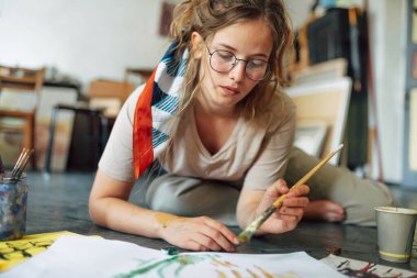 Resim stüdyosunda yerde oturan ve boya fırçasıyla kağıda resim yapan bir kadın sanatçının portresi. Atölyede gözlüklü, suluboya resimli genç bir kadın ressam..