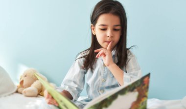 Küçük kız yatakta oturup kitap okuyor ve sessiz bir jest yapıyor. Pijamalı tatlı bir çocuk dinleniyor, işaret parmağıyla dudak işareti yapıyor ve uyumadan önce ilginç hikayeler okuyor.. 