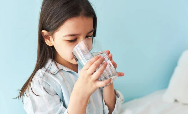 Portrait of a little girl drinking fresh water in the bed at home. Cute preschool kid holding glass of pure water in the morning. Healthy lifestyle concept.