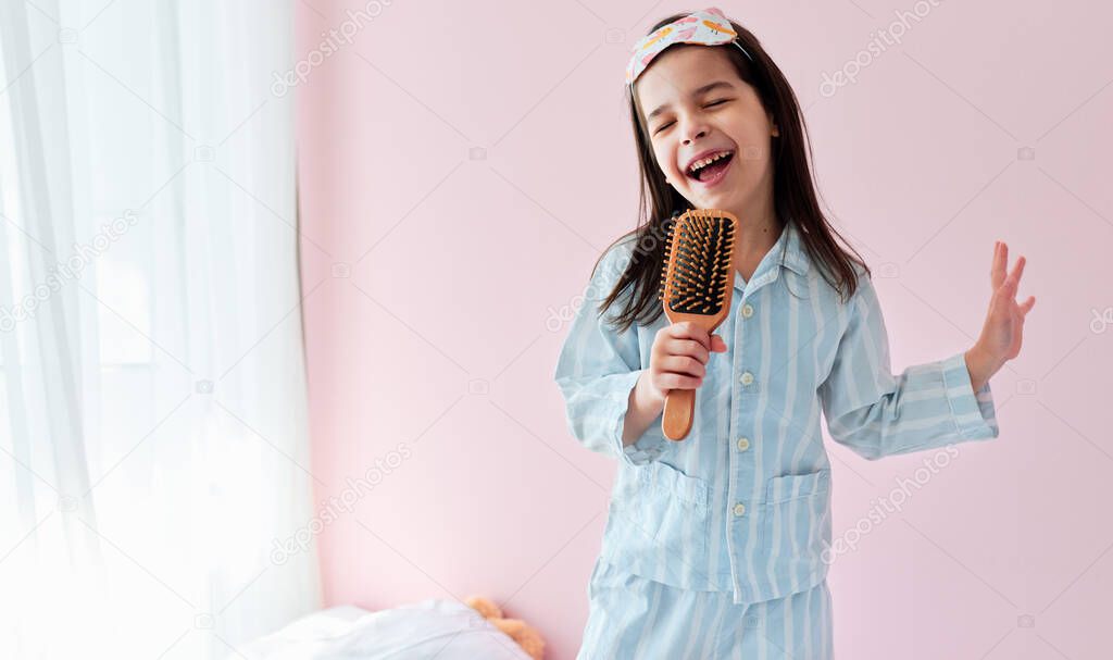 Portrait of a joyful little girl in pajama on the bed holding a hair brush like microphone singing imitates herself a real singer in the morning.