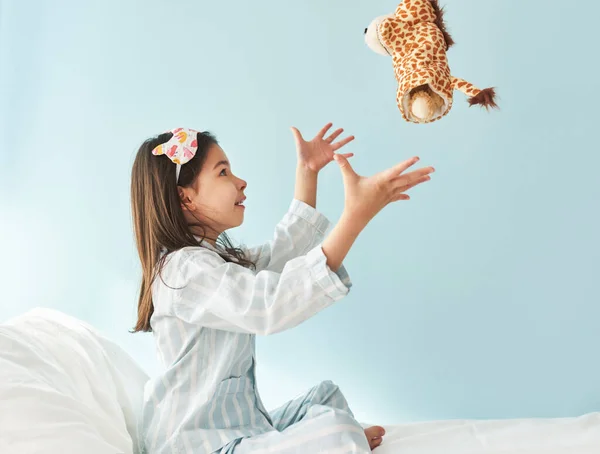 Sied view of cute little girl in blue pajama in white stripes, sleeping mask on head, smiling, and playing with toy giraffe in the bed. Kid has joyful expression in the morning.