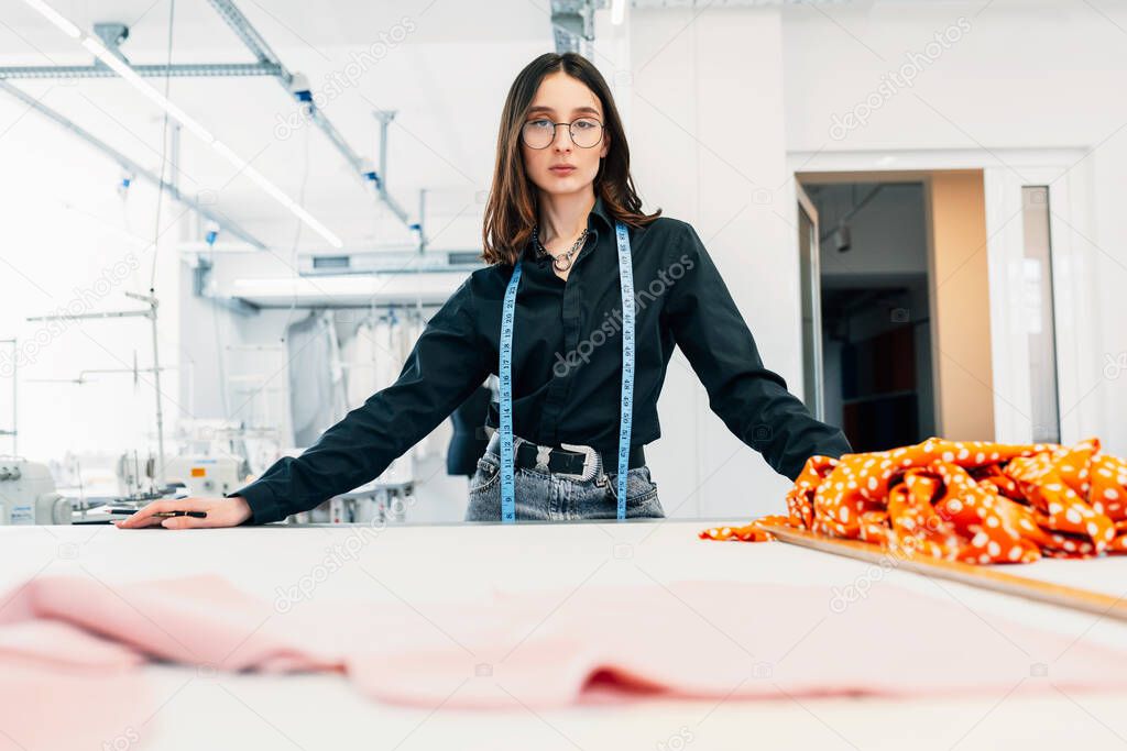 Portrait of a fashion designer looking at the camera posing in her creative workshop. Young woman business owner working with a garment for a new collection in her creative office. 