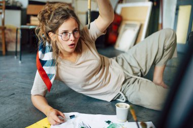 Resim stüdyosunda yerde oturan ve kağıt üzerinde fırçayla resim yapan bir kadın sanatçının yatay görüntüsü. Atölyede suluboya gözlüklü güzel bir kadın ressam..