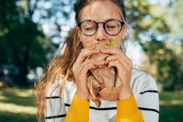 学生の女性のクローズアップ肖像画は 屋外でサンドイッチと昼食を持っています 眼鏡のブロンドの若い女性が公園でファーストフードを食べるために休息を取る — ストック写真