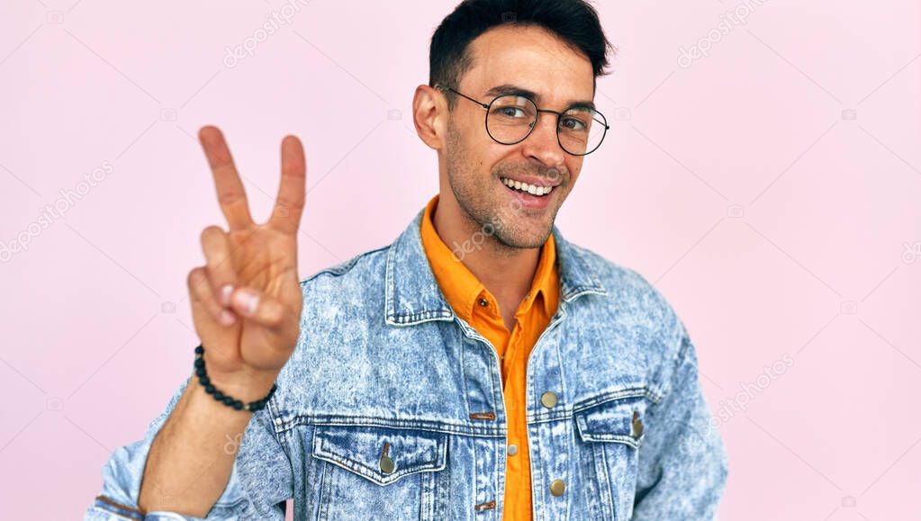 Happy young man in casual stylish clothes, transparent eyeglasses showing victory gesture, isolated on pink studio background. Joyful male making peace gesture.