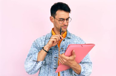 Genç erkek yönetici yeni bir başlangıç yapıyor, odaklanmış ifade ile sözleşme yapıyor, yüzünde kalem tutuyor, pastel pembe arka plan üzerinde izole edilmiş.. 