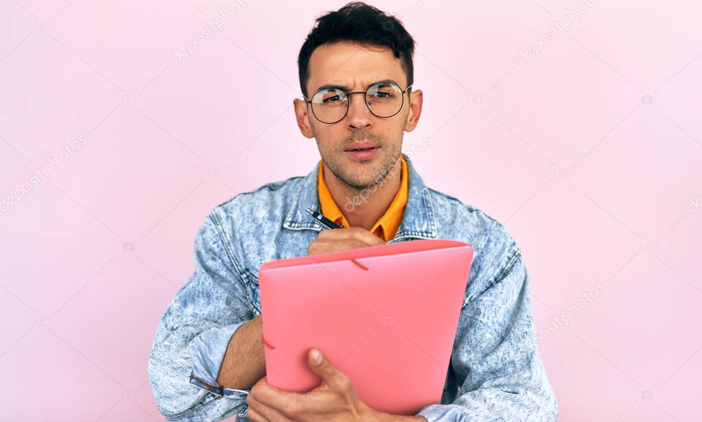 Interested male writes down planning tips in the document with pencil, listens attentively to what to do, isolated over pastel pink background.